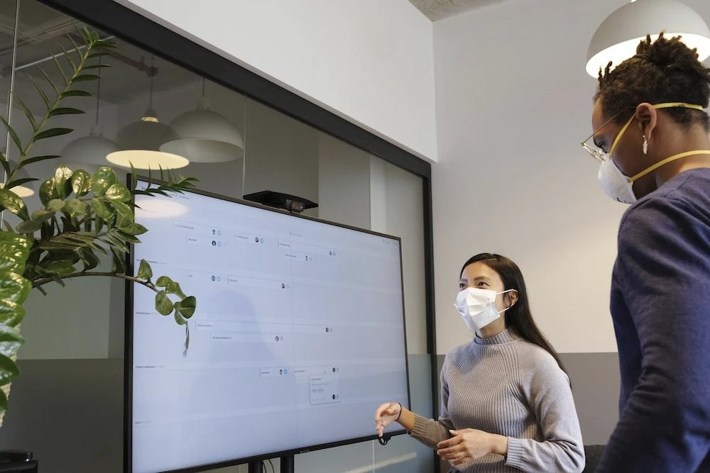 Two colleagues in masks looking at a large wall screen in an office
